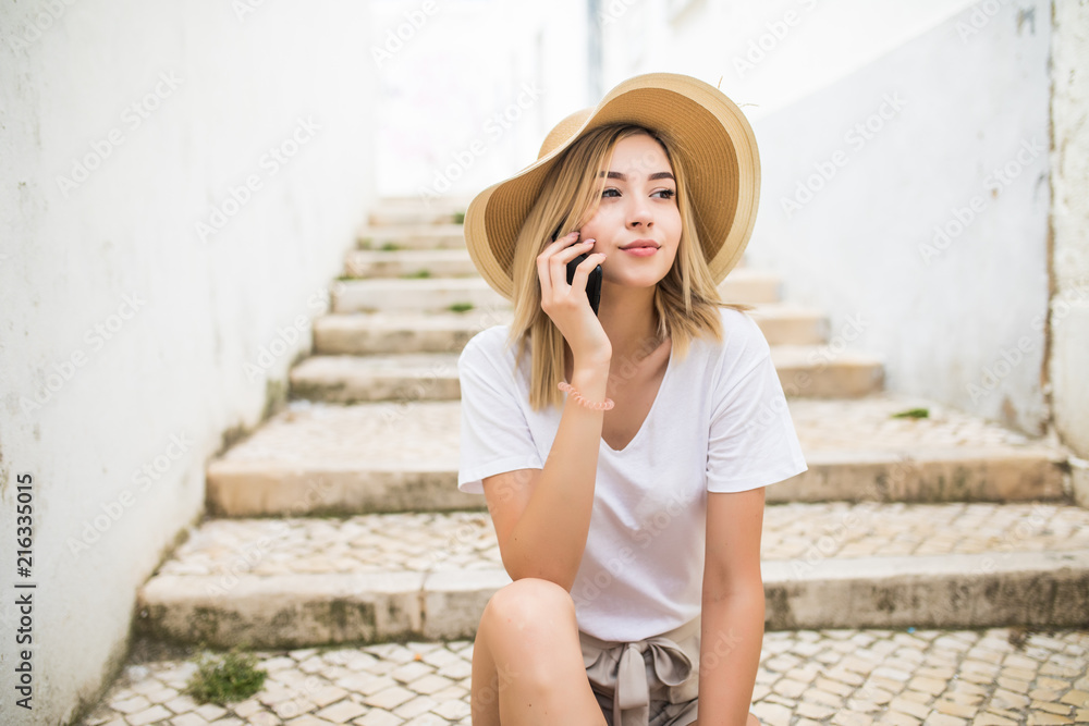 Photo of beautiful young woman sitting on street stairs on summer day and having mobile conversation on smartphone