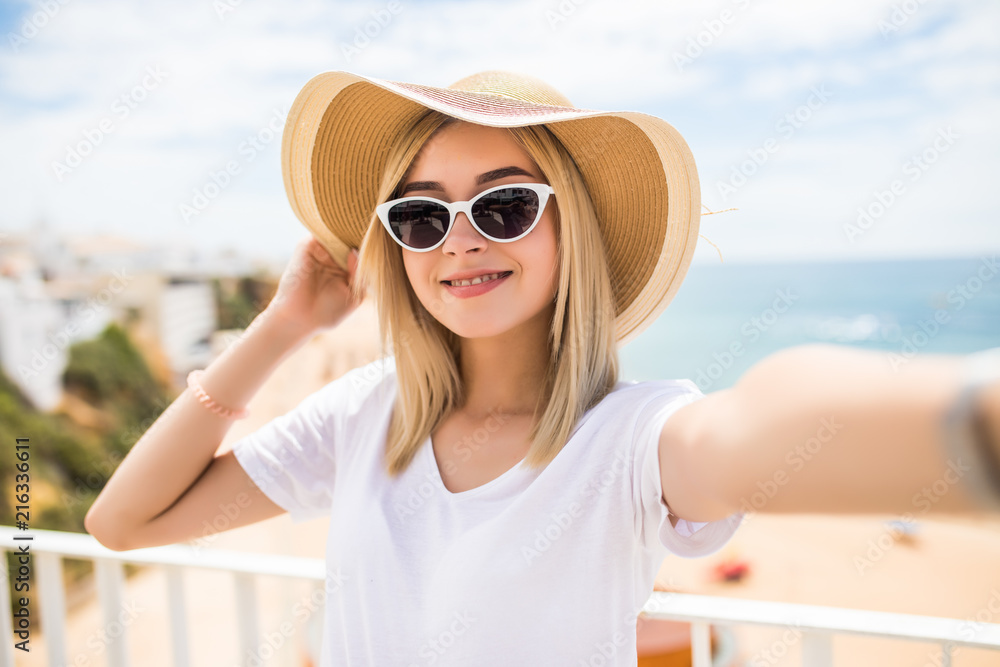 Beautiful woman in sunglasses and summer hat taking selfie on beach