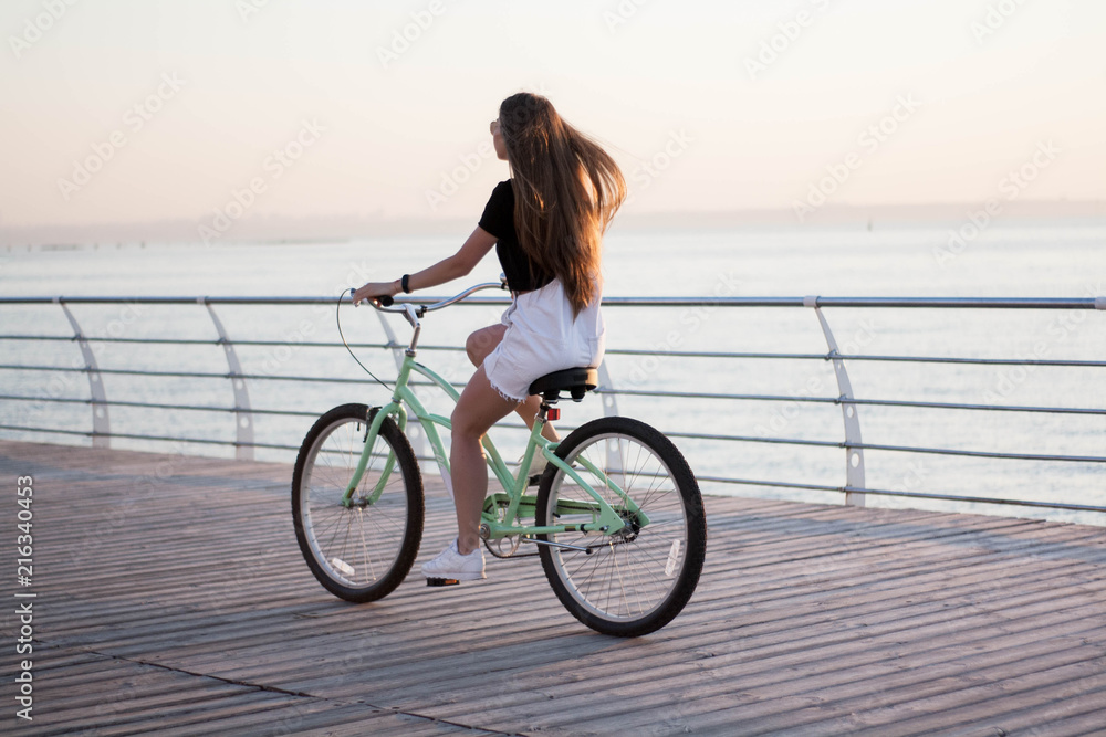 Fototapeta premium Beautiful longhaired young woman with a bicycle having fun, cycling, posing on the camera near the sea on the sunrise
