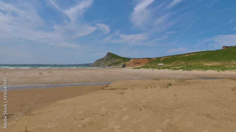 A view of the beautiful wide sandy beach and ocean waves against a bright blue sky with a grassy bluff overlooking the coast. A view of the beautiful wide sandy beach and ocean waves against a bright blue sky with a grassy bluff overlooking the coast.