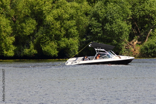 White boat tug boat with tower and black bimini-top awning stands on the water with a sagging halyard on the background of green shore-active rest on the river, water skiing, wakeboard, summer