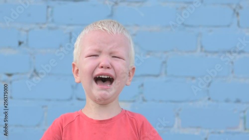 Little blond boy in red T-shirt is crying with tears against gray brick wall.