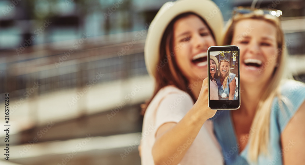 Laughing friends taking selfies together outside on sunny summer