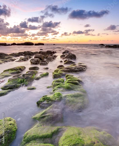Obraz widok pięknego zachodu słońca na plaży z naturalnymi przybrzeżnymi skałami pokrytymi zielonym mchem. miękkie ogniskowanie dzięki efektowi wolnej migawki.