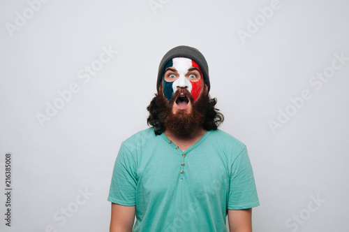 Young bearded man with face painted in french flag colors staring at the camera.