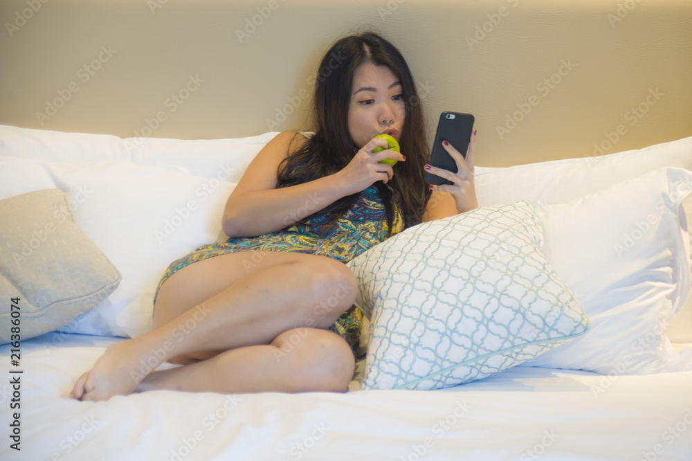 young happy and pretty Asian Korean woman relaxed at home bedroom or hotel room lying on bed eating apple using internet dating texting on mobile phone