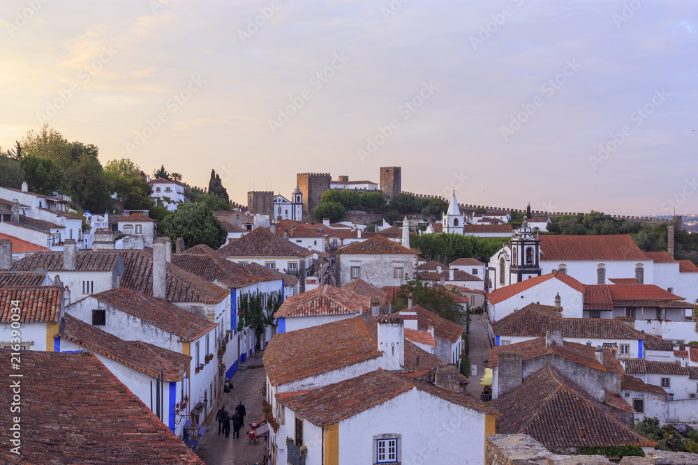 View of narrow streets and castle of Obidos from wall of fortress. Scenic old town with medieval architecture in the sunset. White houses red tiled roofs.