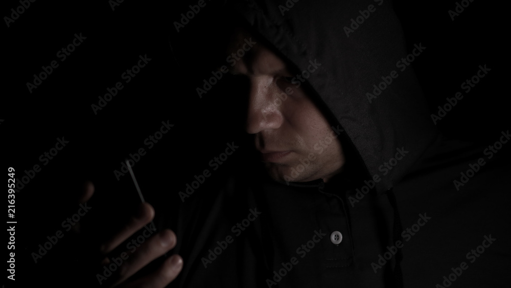 The man's portrait in a hood with the mobile phone in the dark. The man's portrait in a hood with the mobile phone in the dark.