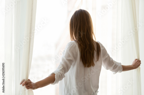 фотографія Back view of millennial woman open white curtains in modern light apartment, hap