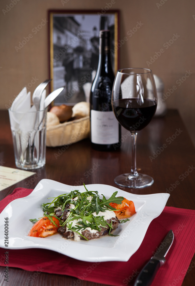 Meat with arugula on a white plate with a glass of red wine in a restaurant