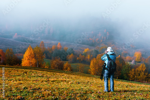 Photographer taking photo of autumn landscape with foggy peaks and orange trees. Ukrainian Carpathians mountains © Ivan Kmit