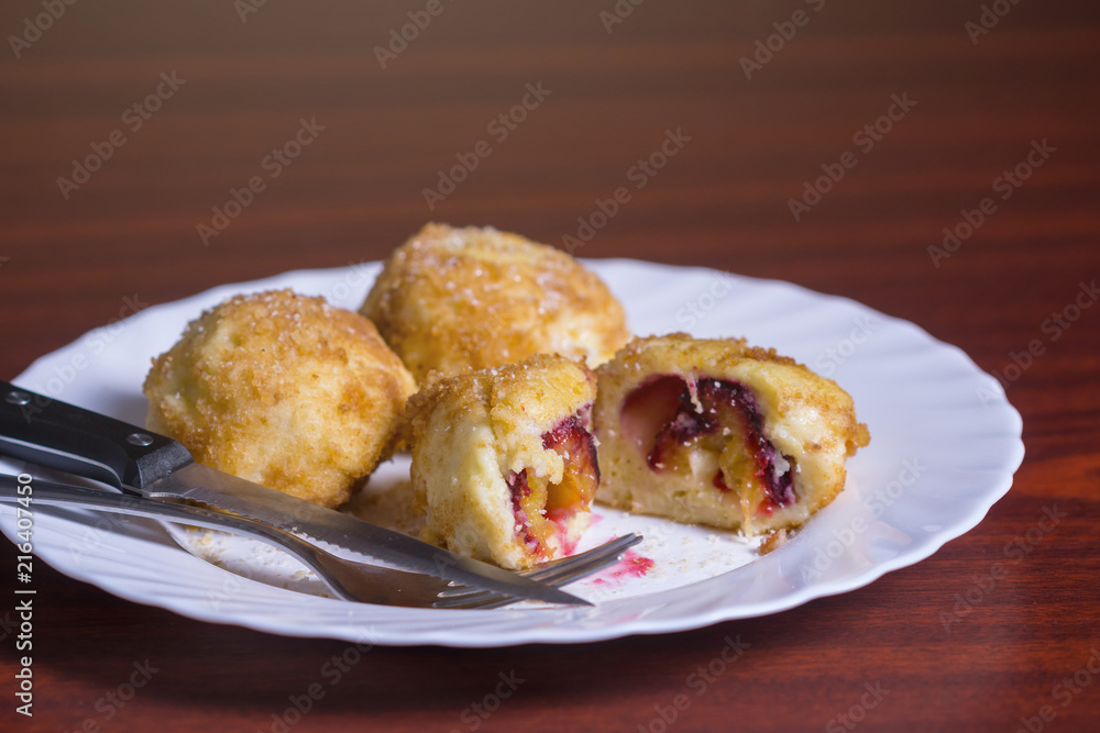 Fresh plum dumplings. One sliced dumpling and two whole with knife and fork on white plate on dark red table Fresh plum dumplings. One sliced dumpling and two whole with knife and fork on white plate on dark red table