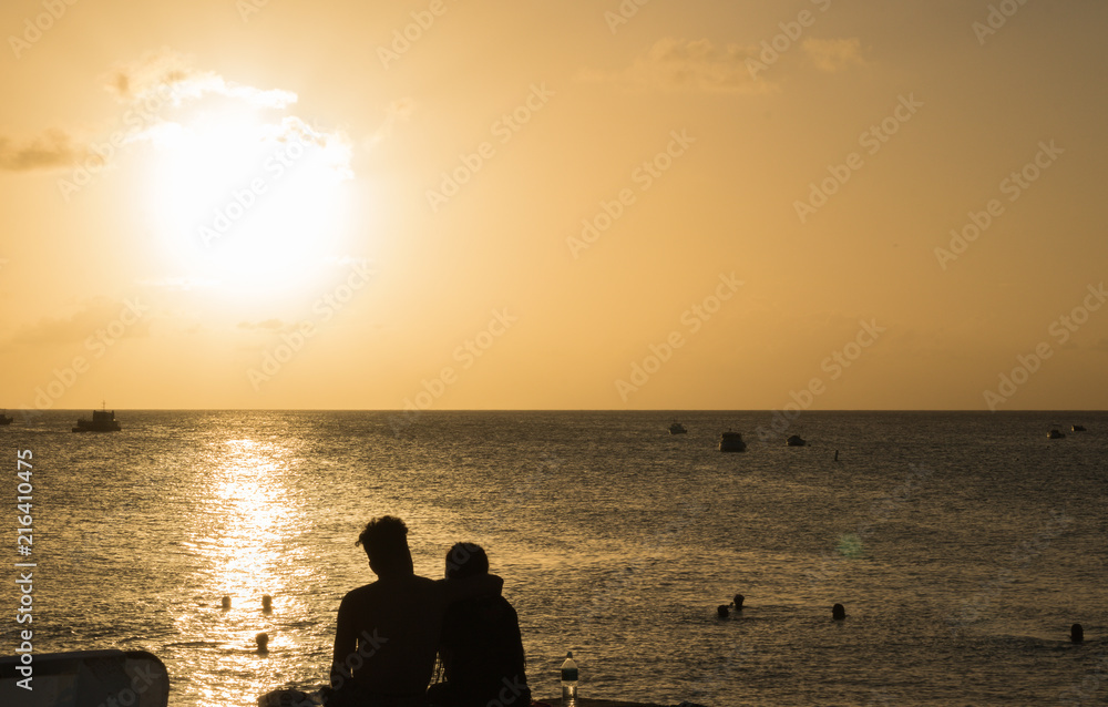 A young couple in love sits in an embrace and looks at the sunset over the horizon of the sea, swimming people and fishing boats in the distance. Silhouettes. Concept: Holiday together.