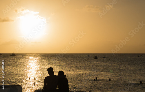A young couple in love sits in an embrace and looks at the sunset over the horizon of the sea, swimming people and fishing boats in the distance. Silhouettes. Concept: Holiday together.