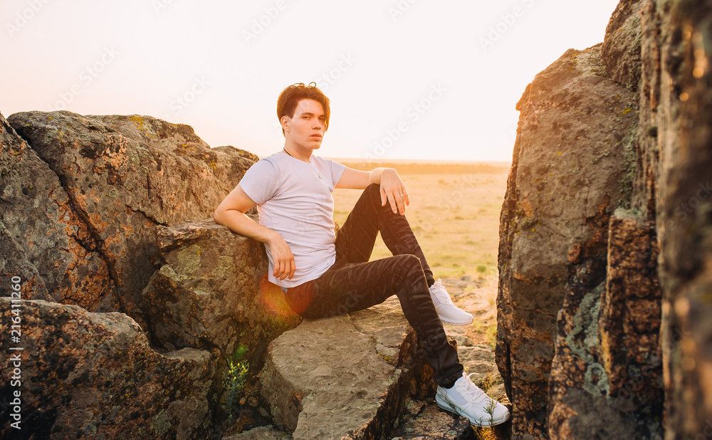 guy sits on top of a mountain between stones in a t-shirt, sneakers and jeans enjoying the sunset guy sits on top of a mountain between stones in a t-shirt, sneakers and jeans enjoying the sunset
