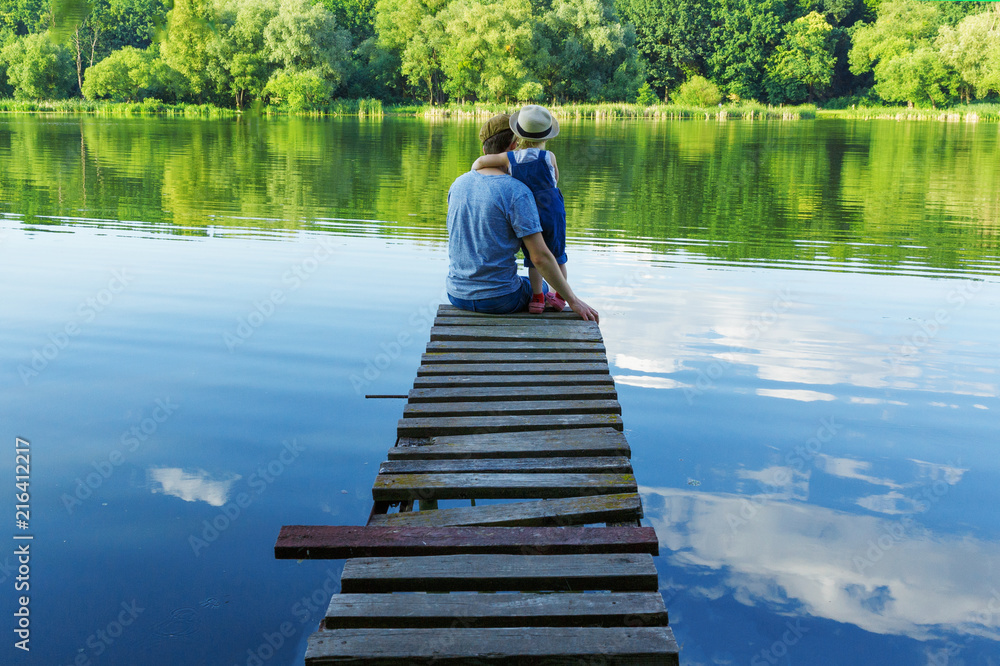 Dad and a tiny girl in dungarees relax at the river. Back view, copy space.