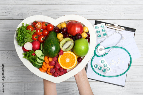 Female doctor holding plate with fresh fruits and vegetables over table, top view. Cardiac diet