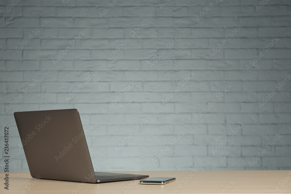 Modern laptop and mobile phone on table against brick wall Modern laptop and mobile phone on table against brick wall