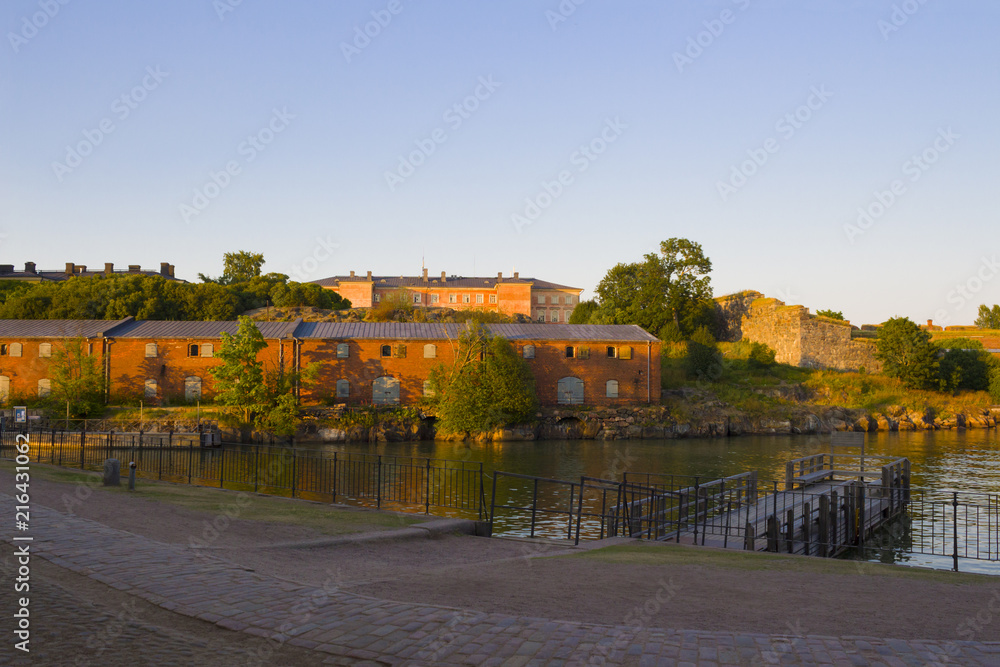 Fototapeta premium A wooden pier and old historical stone fortifications of Fort Suomnelinn in the Gulf of Finland near the city of Helsinki at the end of a summer day.