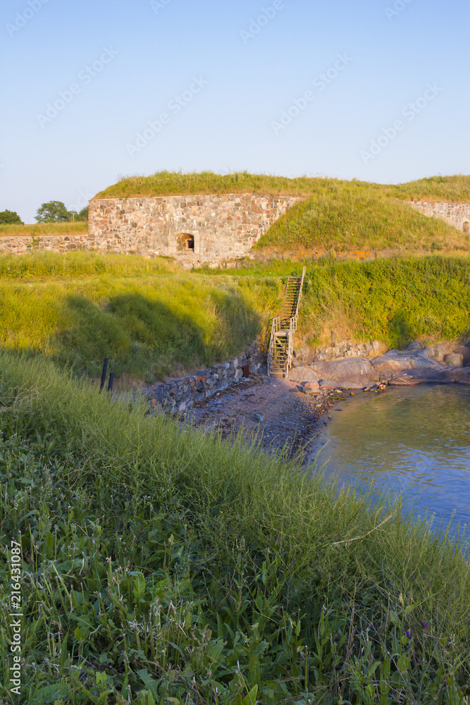 Obraz premium A small cove and old stone fortifications overgrown with grass in the old Fort Suomenlinna in the Gulf of Finland on a summer evening.