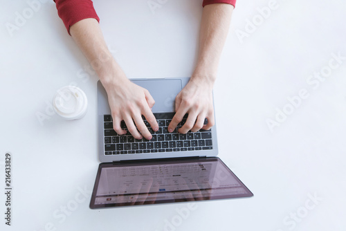 Man typing the text on the laptop keyboard and a white glass of coffee on a light background. Work in the work place. Top view. Copyspace