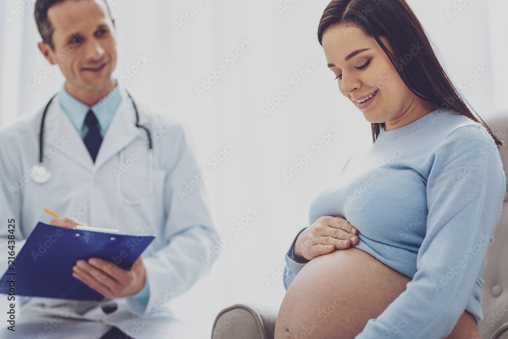 Feeling happiness. Beautiful future mommy bowing her head while watching her belly Feeling happiness. Beautiful future mommy bowing her head while watching her belly