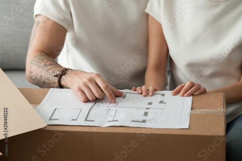 Close up of relocated couple discussing home blueprint on cardboard boxes, considering house remodeling, husband and wife planning apartment interior design, sharing ideas on flat renovation