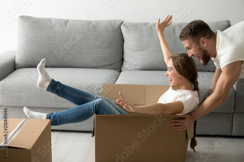 Happy couple play childish riding in cardboard boxes while moving into new apartment together, husband and wife have fun when unpacking cartons relocating to first shared flat. Living together concept photo