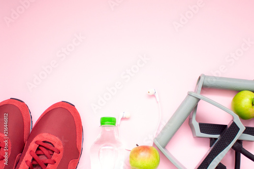 Fitness flat lay. Healthy lifestyle and sport concept. Red sneakers, white earphones, expander, apples and bottle of water on a pink background.