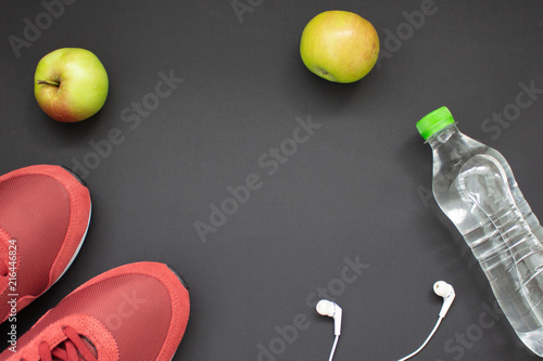 Fitness flat lay. Healthy lifestyle and sport concept. Red sneakers, white earphones, apples and bottle of water on a black background.
