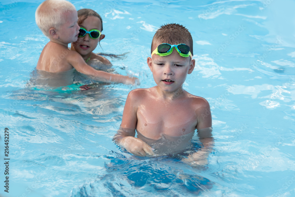 Funny young girl and boys in rainbow glasses for swimming play in blue water of frame pool. Children are happy to swim together in hot summer weather in clear pure water. Сoncept of healthy lifestyle