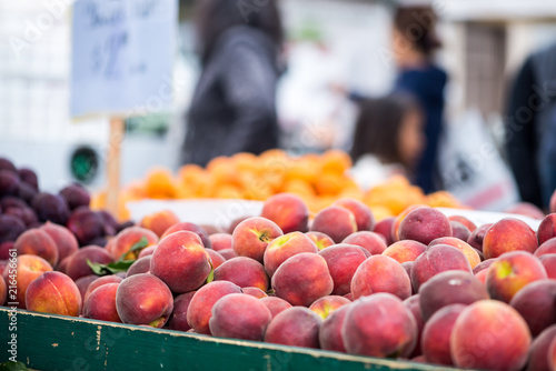 Bin filled with fresh peaches at a farmers market.  Shoppers and signage out of focus in the background.