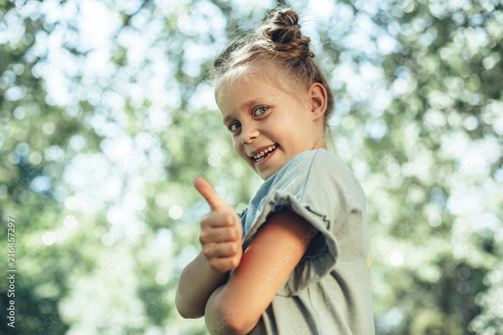 Side view of happy small girl standing in park with folded hands and thumb up. She is smiling at camera with joy Side view of happy small girl standing in park with folded hands and thumb up. She is smiling at camera with joy