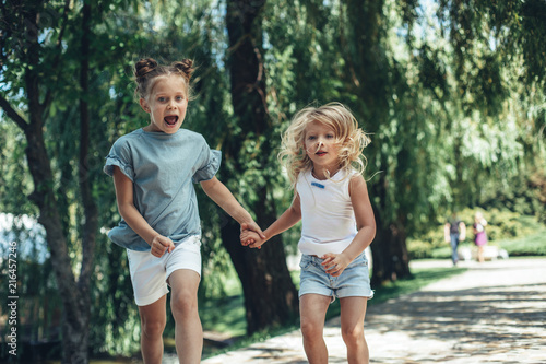 Full length of delighted sisters running in park and holding hands. Older girl is leading younger one and with mouth wide open in content