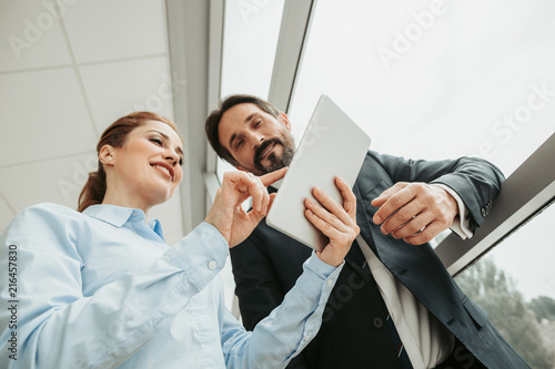 Low angle portrait of cheerful girl typing in electronic tablet while speaking with happy bearded man