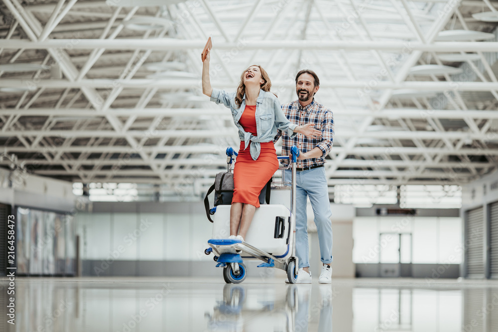 Fototapeta premium Full length portrait of laughing girl and cheerful bearded male having fun in airport. Entertainment of happy couple concept