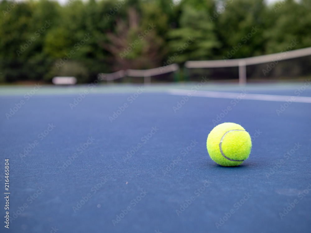 Tennis ball on ground with court nets out of focus. 