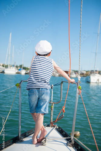 little boy in captain hat standing on white yacht board in sea port in summer