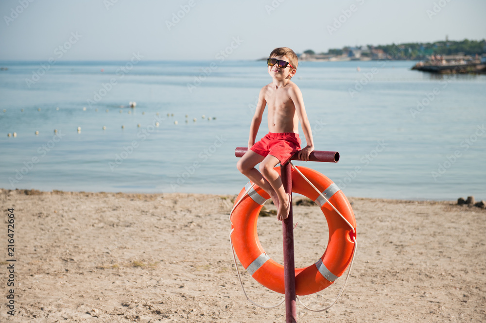 healthy little boy in sunglasses sitting on beach with lifebuoy in summer day healthy little boy in sunglasses sitting on beach with lifebuoy in summer day