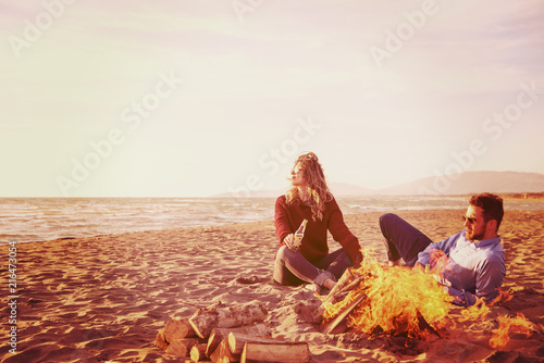 Young Couple Sitting On The Beach beside Campfire drinking beer