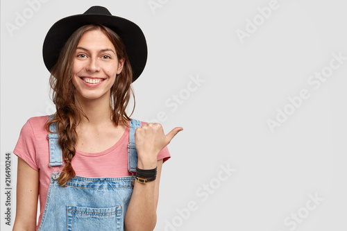 Horizontal shot of positive female ranch owner indicates at her property, shows garden with rich harvest, has happy expression, wears elegant black hat, isolated on white studio wall with blank space