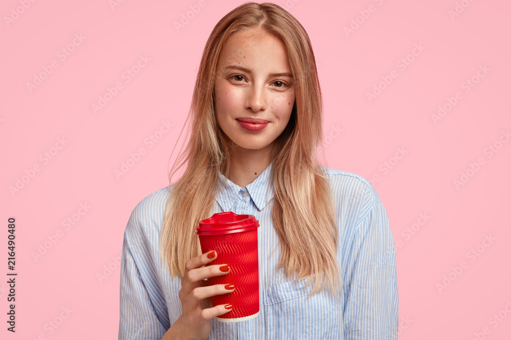 Attractive blonde female student holds hot beverage in disposable paper cup, wears elegant shirt, stands against pink background, has break after having lectures. People and spare time concept Attractive blonde female student holds hot beverage in disposable paper cup, wears elegant shirt, stands against pink background, has break after having lectures. People and spare time concept