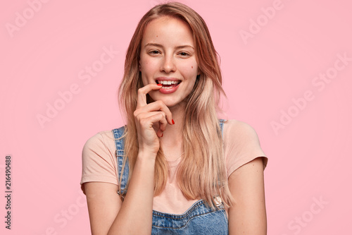 Glad Caucasian female bites fore finger, looks joyfully, has happy expression, being in good mood, dressed in casual clothes, stands against pink background. People, emotions, lifestyle concept