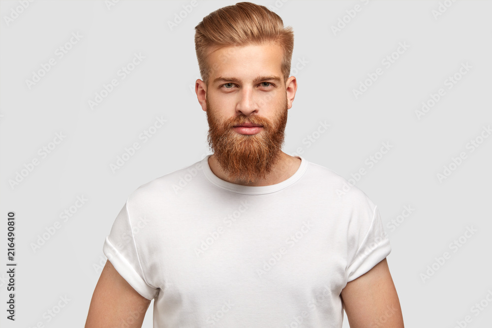 Horizontal shot of prosperous male enterpreneur with appealing look, thick ginger beard, has serious look at camera, listens attentively investors, wears casual t shirt, isolated on white wall Horizontal shot of prosperous male enterpreneur with appealing look, thick ginger beard, has serious look at camera, listens attentively investors, wears casual t shirt, isolated on white wall