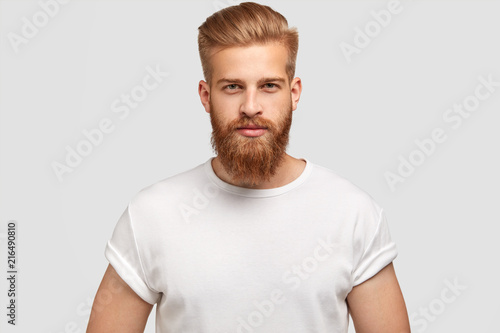 Horizontal shot of prosperous male enterpreneur with appealing look, thick ginger beard, has serious look at camera, listens attentively investors, wears casual t shirt, isolated on white wall