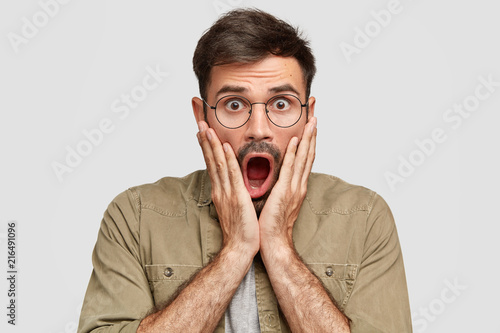 Emotive scared shocked male with widely opened mouth hears impressive rumor, stares at camera with stunned expression, being in stupor, wears round glasses, isolated over white studio background © wayhome.studio 