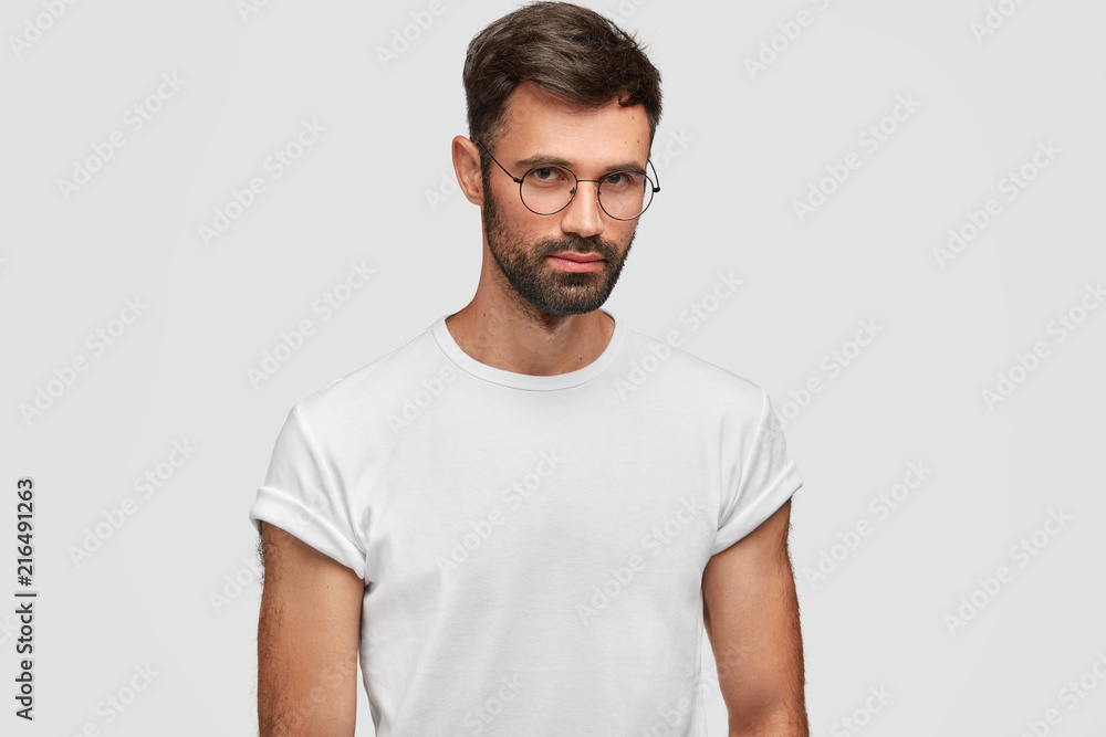 Waist up portrait of handsome bearded male with serious expression, contemplates about something, wears round spectacles and white casual t shirt, poses indoor. People and facial expressions Waist up portrait of handsome bearded male with serious expression, contemplates about something, wears round spectacles and white casual t shirt, poses indoor. People and facial expressions