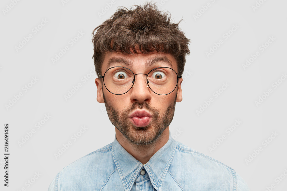 Funny bearded young male rounds lips and has eyes popped out, has comic facial expression, wears round spectacles and denim shirt, expresses disbelief, reacts on something amazing, stands indoor