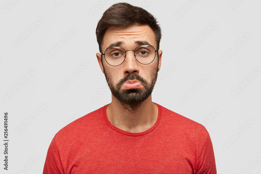 Fototapeta premium Gloomy displeased sorrowful bearded young male purses lips in discontent, being offended by bad comments of followers, expresses negativity, wears red jacket, stands against white studio wall
