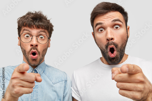 Astonished two bearded guys have surprised expressions, point at camera, have frightened expressions, stand shoulder to shoulder against white background, notice something terrific in distance